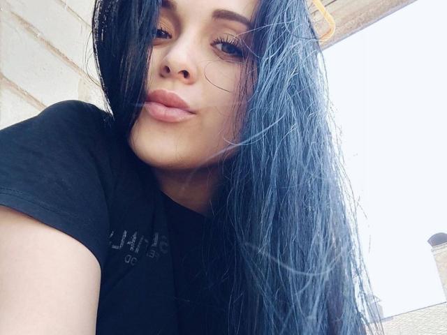 IziPaterson with long black hair, slightly parted lips, and black shirt, leaning against a wall, looking down with a small smirk, bright sunlight on her face.