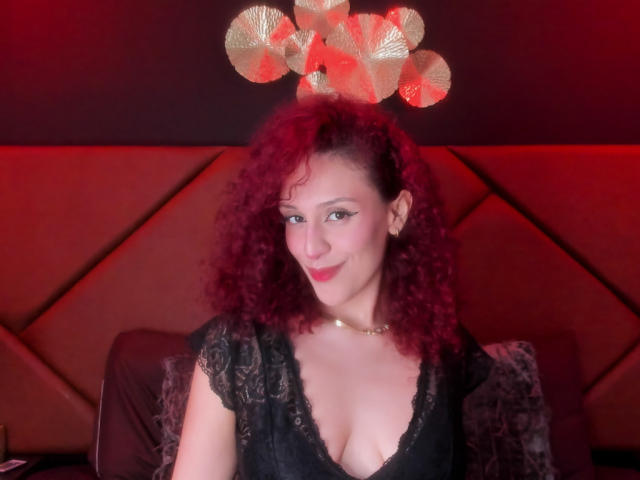 Kaylin Krof, com cabelo ruivo encaracolado, blusa de renda preta e batom vermelho, mostrando os seios, sorrindo, usando um colar de ouro, sob uma luz vermelha com uma coroa de folhas douradas na cabeça..