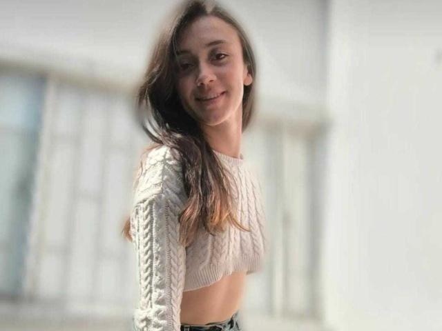 AmyKobold69 with long brown hair, smiling, wearing a white, long-sleeve, knit crop top, exposing her small tits, in front of a blurred, white background.