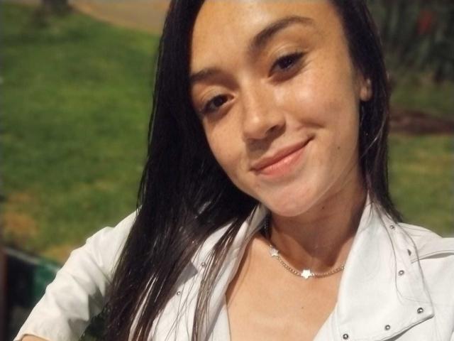 AmyKobold69 has medium brown skin, long black hair, and a subtle smile. She's wearing a white shirt with a small flower necklace. Boobs are not visible.