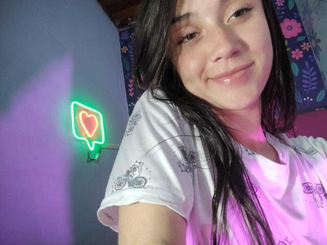 AmyKobold69 with long black hair, white shirt, subtle smile, pinkish nose, neon heart wall light, purple and blue background, and soft pink light on her right.