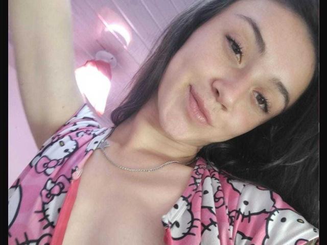 AmyKobold69 with brown hair, smiling, wearing pink Hello Kitty pajama top, showing cleavage, silver necklace, arm raised, selfie, looking at camera, slightly flushed, casual, confident.