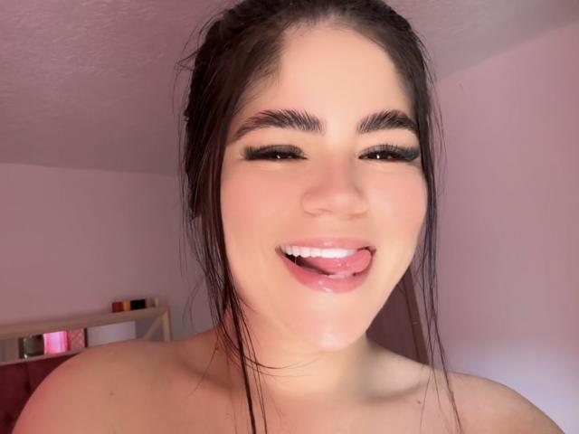 MartinaRossyI with dark hair, thick eyebrows, long lashes, and a playful tongue-out expression, has a shiny, wet pussy on her chin. Her skin is fair and she's topless.