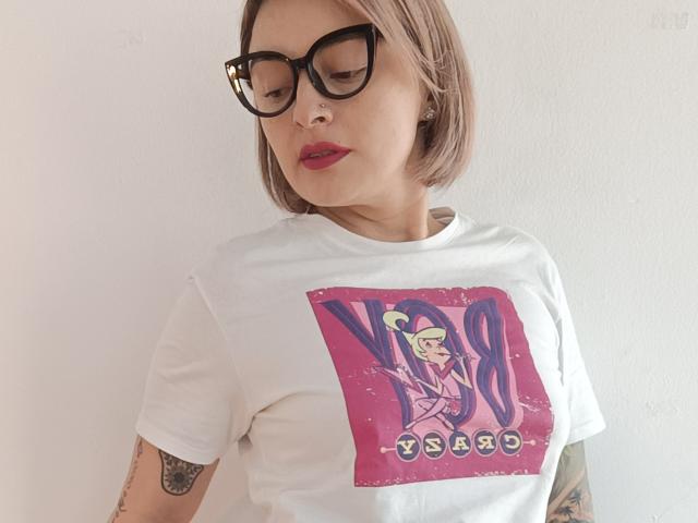 KatParadise with short blonde hair, black glasses, red lipstick, white t-shirt with pink and purple graphic, tattoos on arms, looking down with a serious expression.