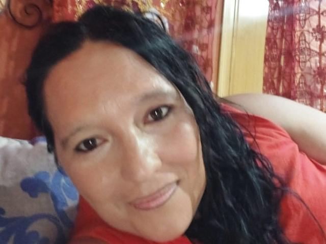 PeteAndJade, a woman with long black wavy hair, light skin, brown eyes, and a slight smile, wearing a red shirt, lying down with a relaxed expression.