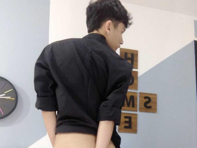 HarryClaude, standing with back to camera, wearing black shirt, no pants, exposing bare ass. He's a young Asian guy with short black hair, looking to the right.