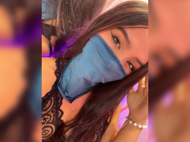 EmyXCute with long dark hair, wearing a blue face mask and black lace top, has her hand in her hair, showing a pearl bracelet on her wrist.