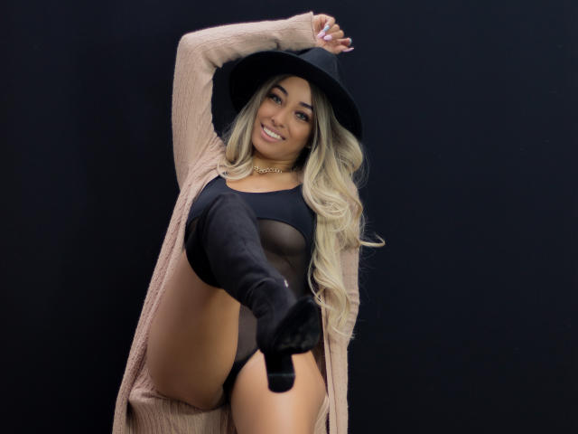 Diana Hopson, con lunghi capelli biondi, cappello nero, top in rete nera e stivali neri che arrivano all'altezza delle cosce, sorride, sollevando una gamba, mostrando la sua vulva scoperta e la pelle abbronzata..
