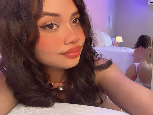 LiliMays with wavy black hair, full pink lips, and winged eyeliner, wearing a pink collar with a bell, lying on a bed, showing her ass in white thong.