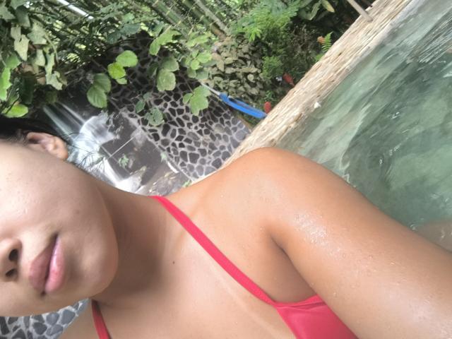 Heyly est dans un bikini rouge, des gouttelettes d'eau sur sa peau bronzée, debout dans une piscine avec une végétation luxuriante à proximité.