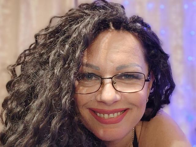 AngellaFly with curly black hair, glasses, red lipstick, smiling at the camera, showing her tits, has a light brown skin tone, and is wearing a necklace.
