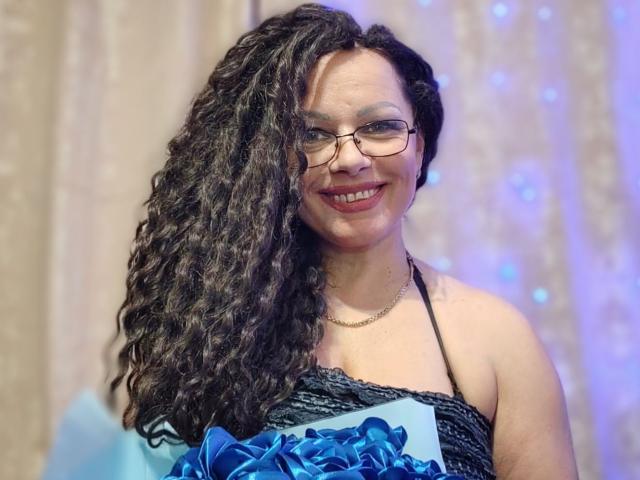 AngellaFly with long curly black hair, glasses, and a smiling face, wearing a black halter dress with blue floral accents, showing her tits and a cute ass.