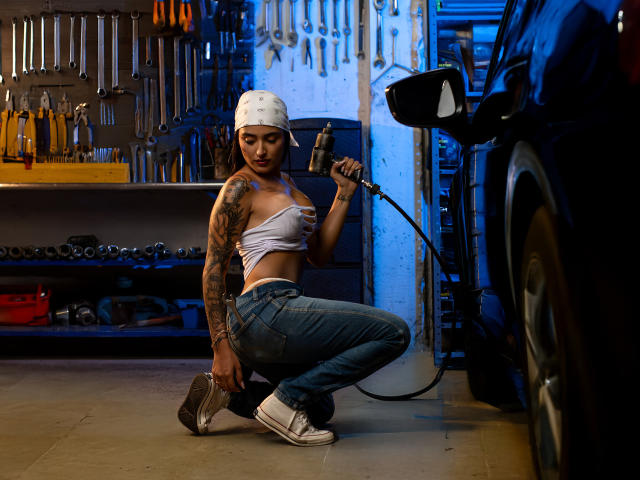 MiaSFoxy, crouching, holding a air hose, white tank top, tattoos, white cap, blue jeans, white sneakers, black car, tool wall, gas station, sexy, large tits, butt.