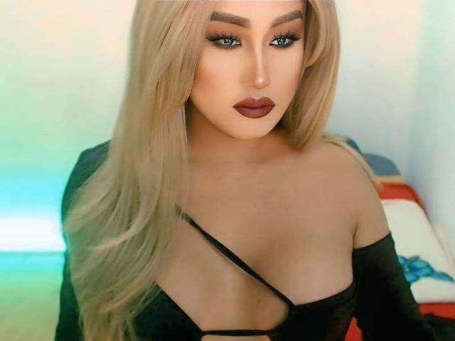 GoddessBellaX with long blonde hair, dark red lipstick, and black strap dress showing her big tits, looking seductive with intense eyes. Soft lighting, sexy, confident.
