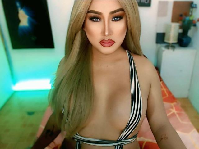 GoddessBellaX with long blonde hair, blue eyes, and red lipstick, wearing a black-and-white striped top, exposing her large tits. She's sitting on a bed with an orange blanket.