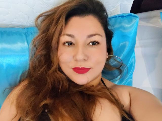 SakotoDika, con lunghi capelli castani, rossetto rosso e orecchini a cerchio, sdraiata su un cuscino blu, che mostra le spalle e il petto scoperti, guarda la fotocamera con un leggero sorriso..
