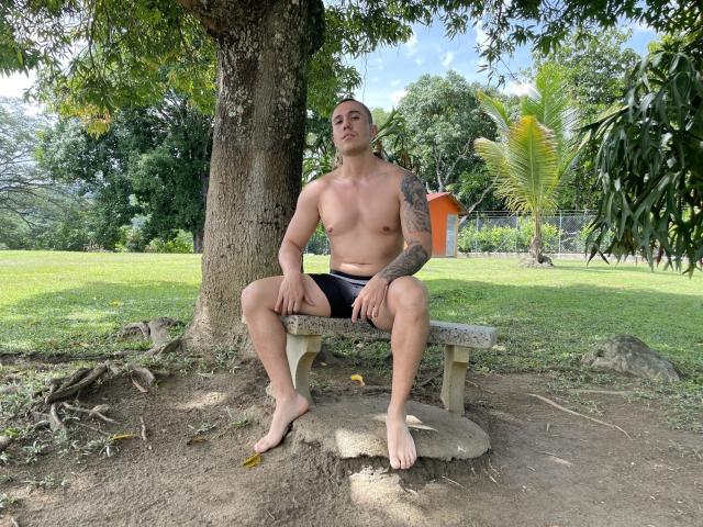 MrDominux, shirtless with tattoos on his right arm, sits on a stone bench under a tree, wearing black shorts, with a neutral expression, in a grassy park.