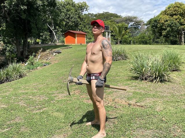 MrDominux stands shirtless in grassy yard, holding axe, wearing red cap, black gloves, and "Dick's" underwear, tattooed arm, muscular, smiling, sunlit, greenery, orange shed in background.