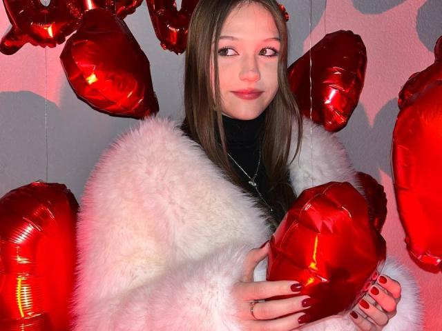 Bubochka with long brown hair, wearing a white fluffy coat, holding red heart-shaped balloons, and sporting red nail polish, smiling at the camera.