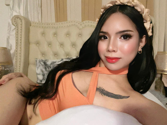 AyeshaSebastian with long black hair, orange halter top, pink lipstick, feather tattoo on chest, beige headband, laying on bed, smiling, showing her tits, holding a pillow.