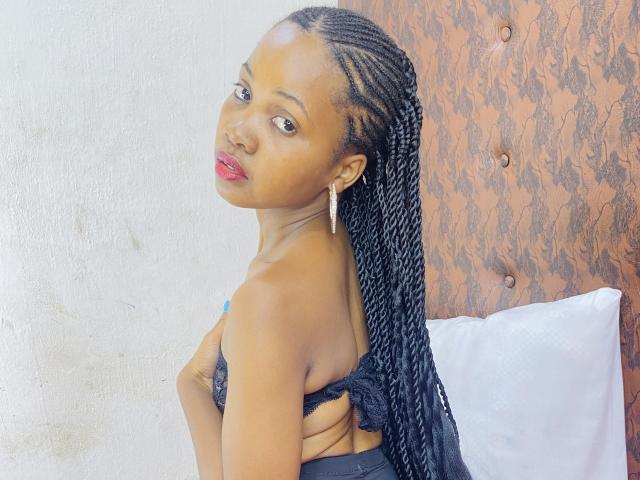 DareMagik with long black braids, red lips, and black lace top, showing her back and looking over her shoulder, wearing a silver earring, standing in front of a pillow.