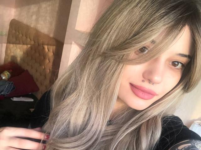 Flimina mit langem, blondem und graumeliertem Haar, rosafarbenen Lippen, Nasenring und leichtem Lächeln, trägt ein schwarzes Oberteil und hält ihre Haare mit roten Nägeln fest, im weichen Sonnenlicht..