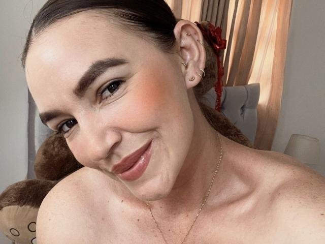 SophieGlamoursX, topless with bare shoulders, smiling with pink blush, dark eyebrows, small nose, ear piercings, and brown hair in a ponytail, wearing a gold necklace.