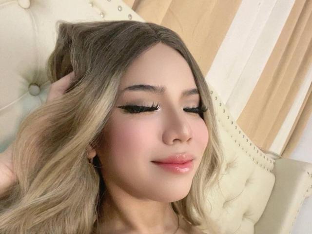AyeshaSebastian has long, wavy blonde hair, glossy pink lips, and thick black eyelashes. She's sitting on a tufted beige chair, smiling with eyes closed, and holding her hair.