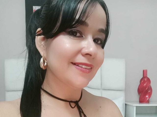 TamaraFoxy with black hair, brown eyes, and pink lipstick, wearing gold hoop earrings and a black choker, smiling slightly, showing her shiny, smooth skin and small, cute nose.