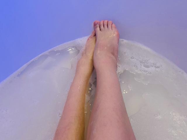 PrimeS's legs in a bathtub, water up to ankles, soles touching, slight bubbles, skin pale, toes pointed upwards, blue-tinted water.