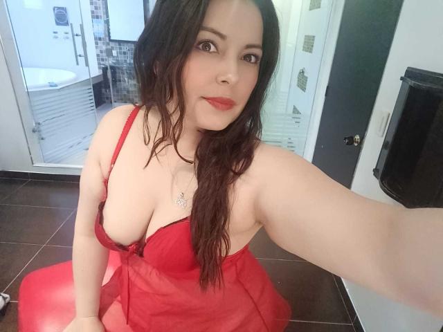 Seher, com o cabelo castanho molhado, usando lingerie vermelha que mostra seus seios grandes, sentada em uma cadeira vermelha, tirando uma selfie em um banheiro. Batom vermelho, sorriso discreto, com o braço estendido..