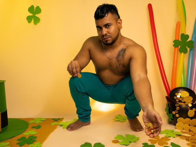 BrianMarly, shirtless, muscular, brown skin, short black hair, crouching, green pants, holding gold coin, silver necklace, tattoos on chest, colorful sticks, green and gold paper, confident expression.