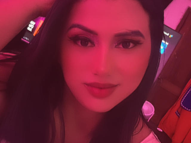 LolaOberle with pink-lit face, dark hair, eyeliner, and slightly parted lips, showing a subtle smile, her hand resting behind her head. Her tits are not visible.