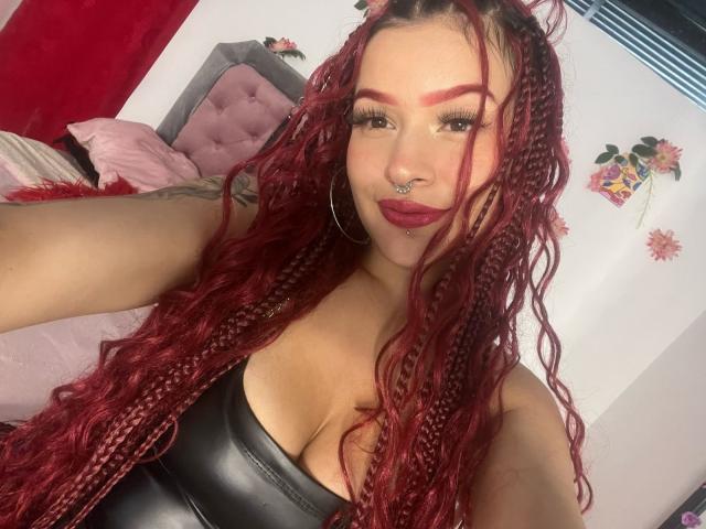 CamiHalli with red braided hair, nose ring, hoop earrings, and dark red lipstick, showing her large boobs in a black leather top, smiling at the camera.