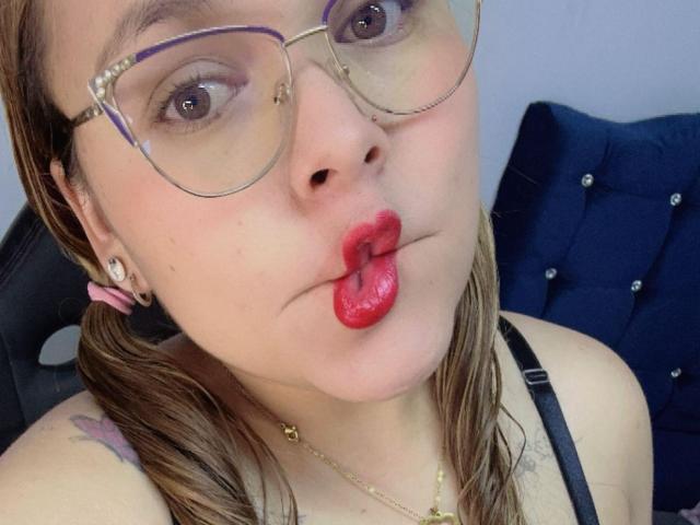 PinkyHotLover with glasses, pouty red lips, and tongue out, has a tattoo on her shoulder, wearing a black strap, and a pink hair clip.