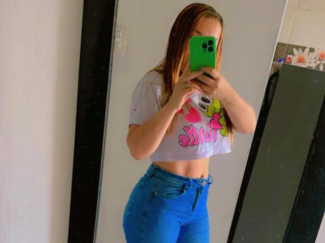 PinkyHotLover mit langen braunen Haaren, die ein weißes Crop-Top mit rosa und gelben Motiven und blaue Jeans trägt, macht ein Selfie vor einem Spiegel mit einem grünen Handy..