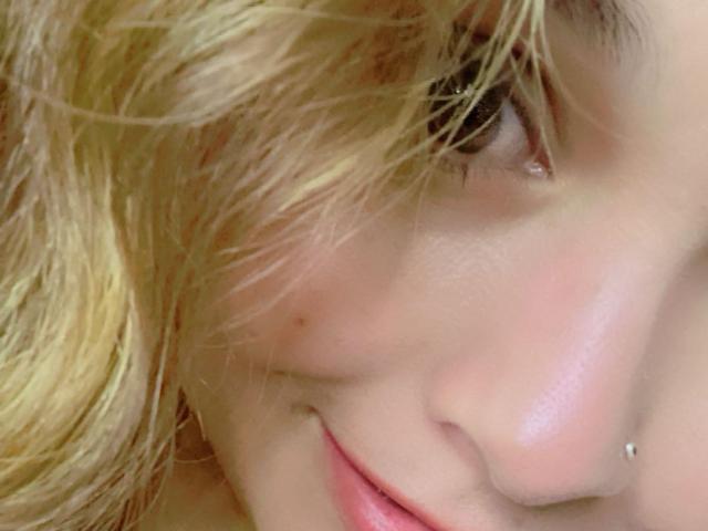 Le visage de PinkyHotLover, avec des cheveux blonds et ondulés qui couvrent un œil, des lèvres roses et un piercing argenté au nez sur le côté droit. Une peau claire et douce avec une petite tache de beauté..