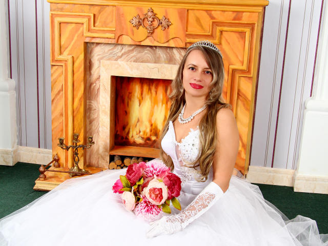 RebeccaDarling69 with long brown hair, wearing a white lace wedding dress, pearl necklace, and silver tiara, holding pink and white roses, sitting on green carpet in front of fireplace.