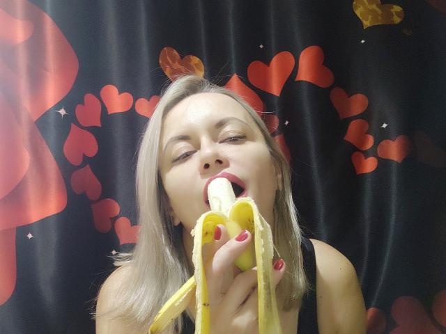 RebeccaDarling69 with blonde hair, red nails, and red bow eating a banana with her mouth open, black sleeve, red hearts in background, looking aroused, banana peel in hand.