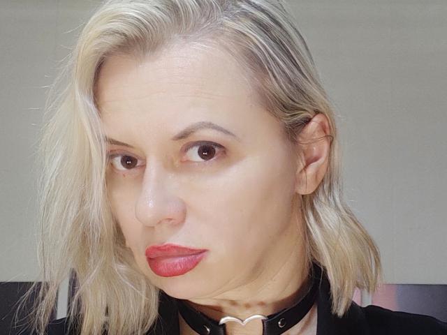 RebeccaDarling69 has blonde hair, red lipstick, a black choker with a heart, and brown eyes. Her face shows slight wrinkles and a neutral expression.