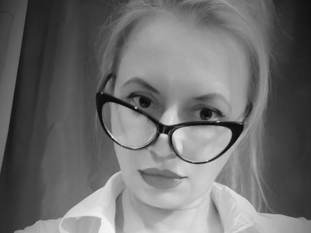 RebeccaDarling69 with big ass glasses, looking straight at the camera, wearing a white shirt, and having light-colored hair in a ponytail, in a black and white photo.