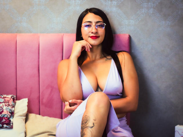 ZaraBeicker with large tits, wearing a low-cut white dress, sitting on a pink bed, hand on chin, glasses, red lipstick, tattoo on thigh, black hair, smiling.
