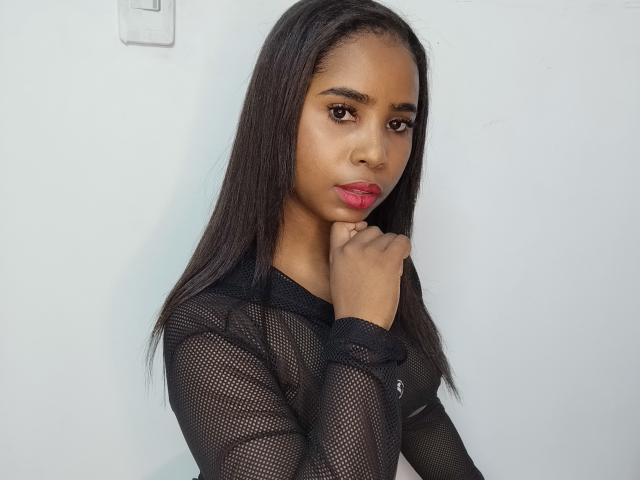LiaLuz with dark, straight hair, brown eyes, and pink lipstick, wearing a black mesh top, touches her chin, looking at the camera, showing her small, cute tits.