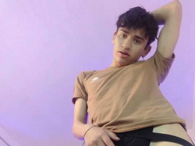 HarryClaude, with curly dark hair, wearing a brown tee, black underwear, and a slightly raised shirt, has a relaxed, slightly provocative, upward-angled pose.