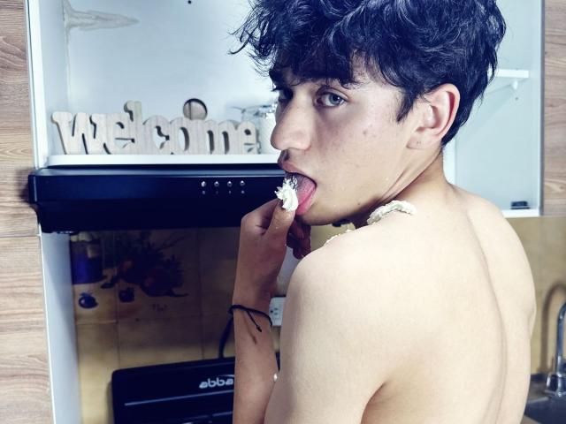 Shirtless HarryClaude with curly black hair, fair skin, and a slim build, licking a white flower with his right finger, looking over his shoulder, in a kitchen.