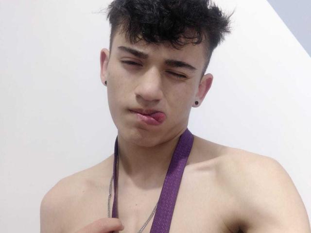 HarryClaude with black hair, black ear studs, and a purple strap, sticking out his tongue, looking slightly annoyed, shirtless with a slim, toned upper body.