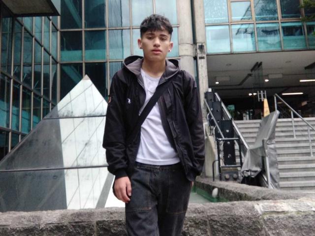 HarryClaude stands with a neutral expression, wearing a black jacket, white tee, and dark jeans, a crossbody strap, and white earbuds, in front of a modern building.