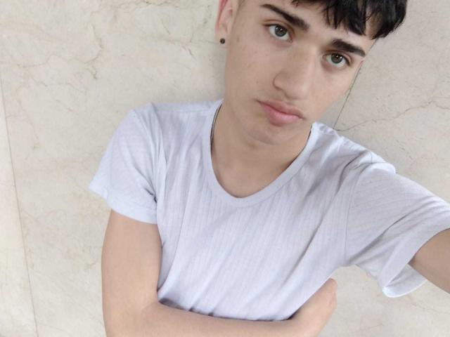 HarryClaude with short dark hair, green eyes, and a slight pout, wearing a white t-shirt, arms crossed, looking up at the camera, against a light marble wall.