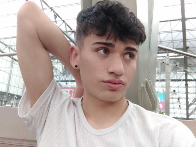 HarryClaude with short, curly black hair, light skin, small black ear stud, wearing a white shirt, arm raised, looking to the side, slightly pouty lips, indoor setting.