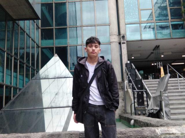 HarryClaude has short dark hair, a serious face, wearing a black jacket over a white tee, and dark pants with a black strap across his chest.